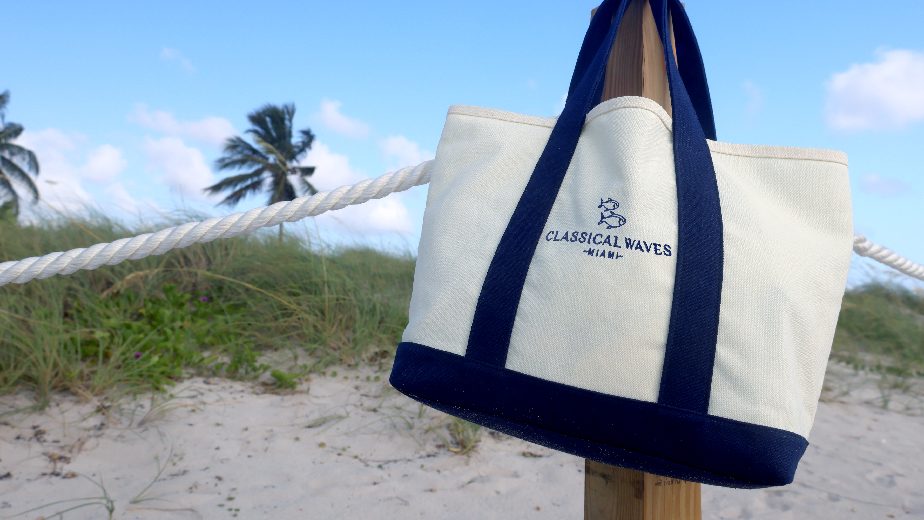 An off-white canvas tote bag with navy blue handles and trim, featuring the 'Classical Vates' logo, hanging from a wooden post on a beach with dunes, palm trees, and a blue sky in the background.